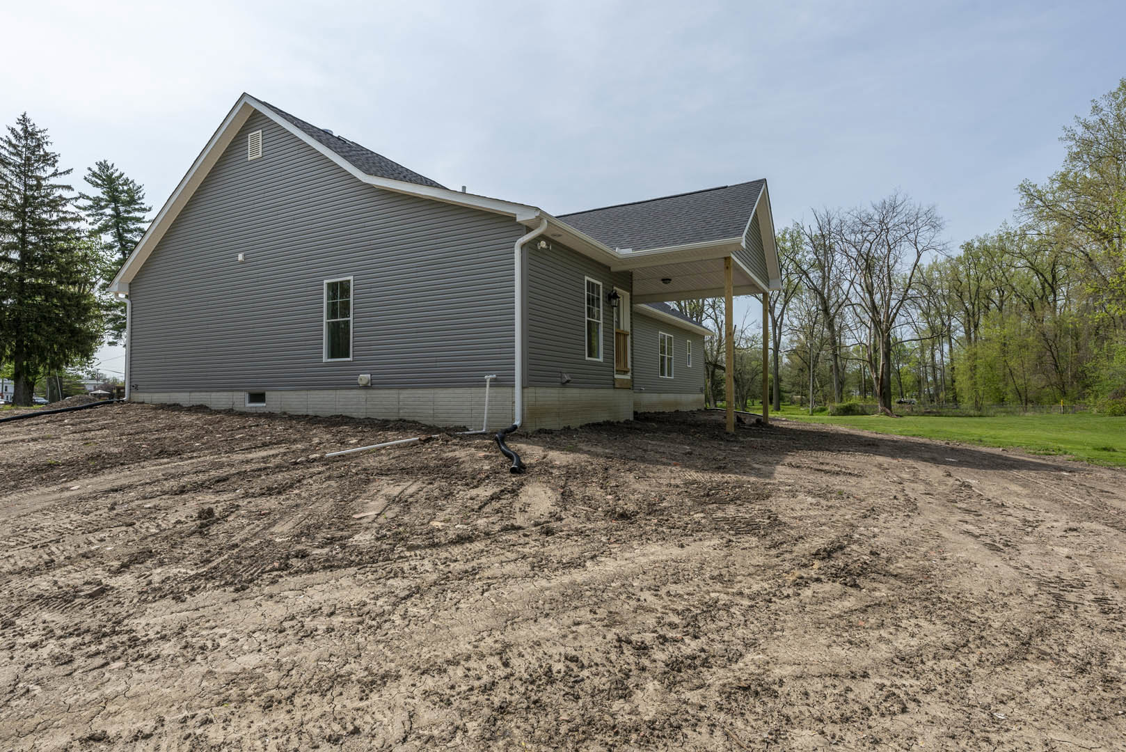 Single-story home with gray roof and white-framed windows, surrounded by leafy trees and dirt ground, black pipe visible along a dirt road, forested area in background.