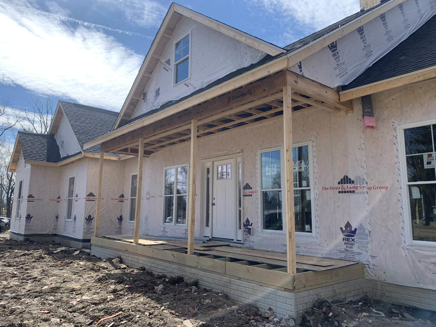 Partially built home with exposed wooden framing, covered porch, white door with glass window, multiple white-framed windows, gray siding, and unfinished roof under cloudy sky.