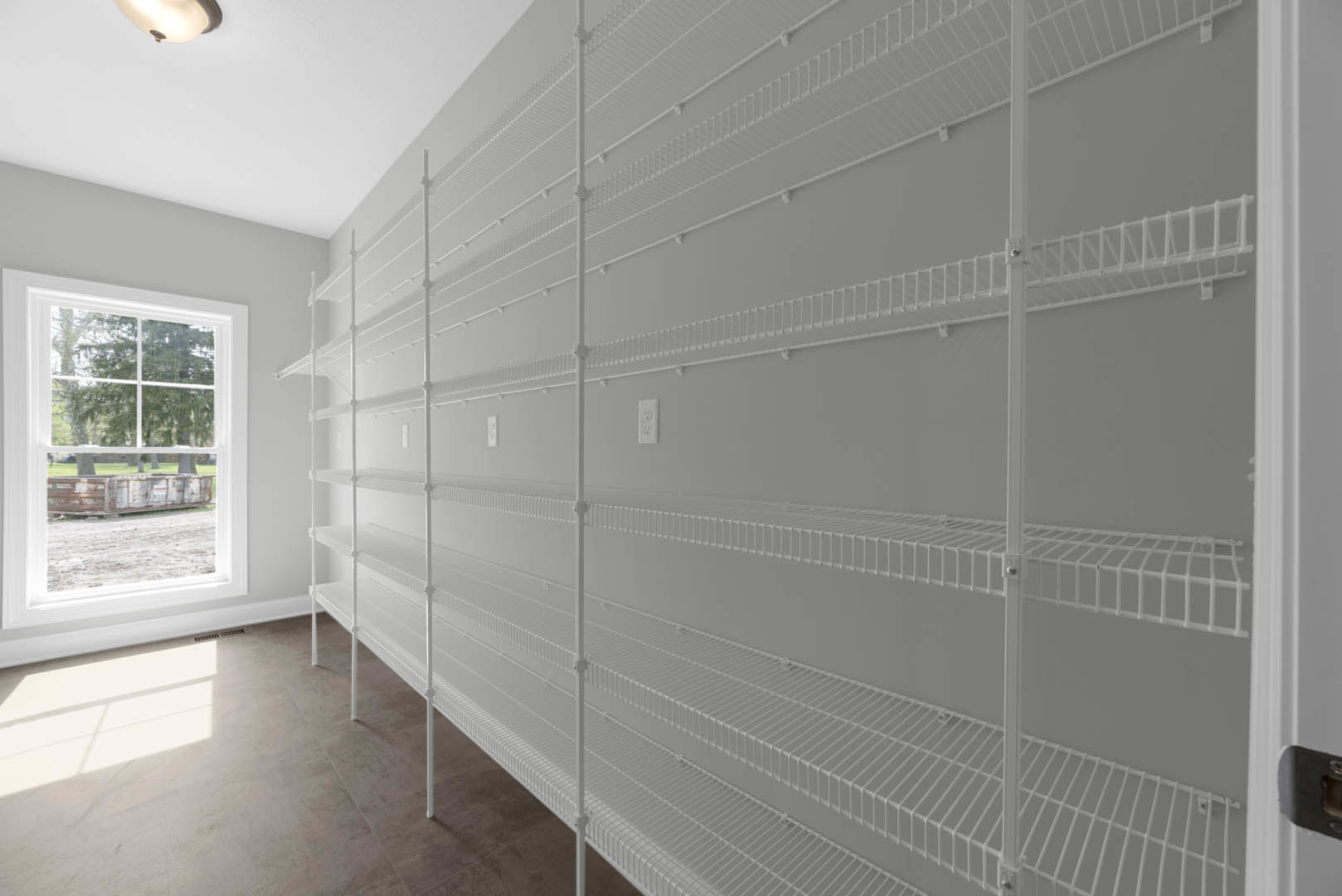 Built-in white shelving on a plaster wall beside a window with a white frame, tile flooring, and ceiling detail; window overlooks a dumpster.