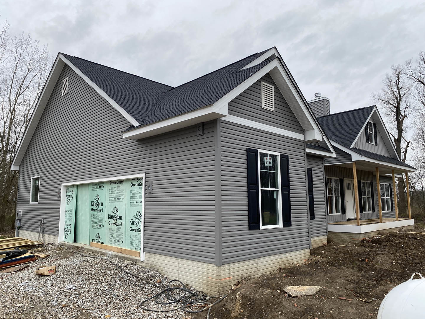 Framed house under construction with exposed wood, white siding panels, and windows, set on a hillside under a cloudy sky
