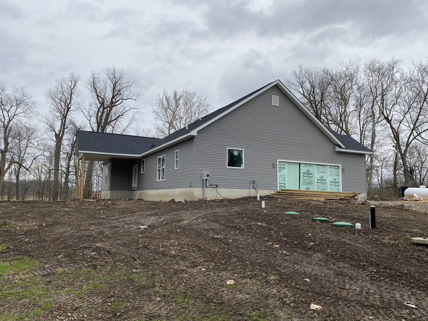 Grey house under construction with white door, window displaying a sign, dirt lot bordered by metal fence, surrounded by trees and cloudy sky