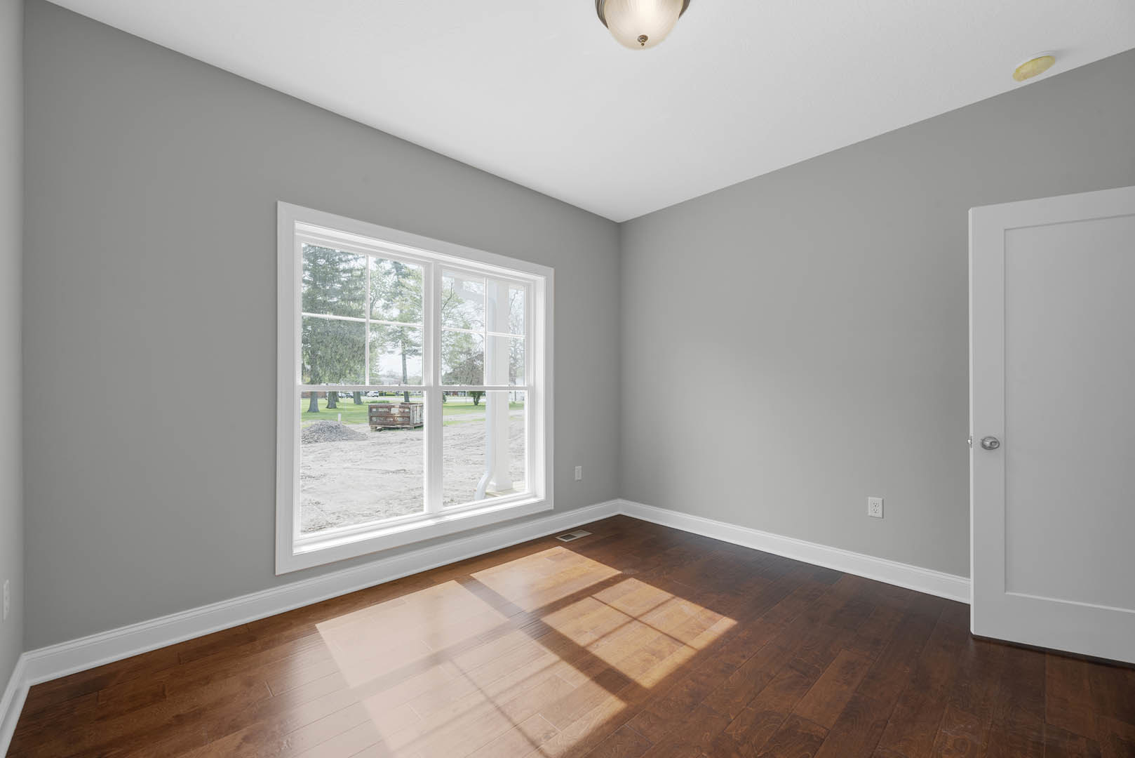 Sunlit room featuring wide-plank hardwood flooring, large window overlooking green yard, white walls with crown molding, and a paneled door.