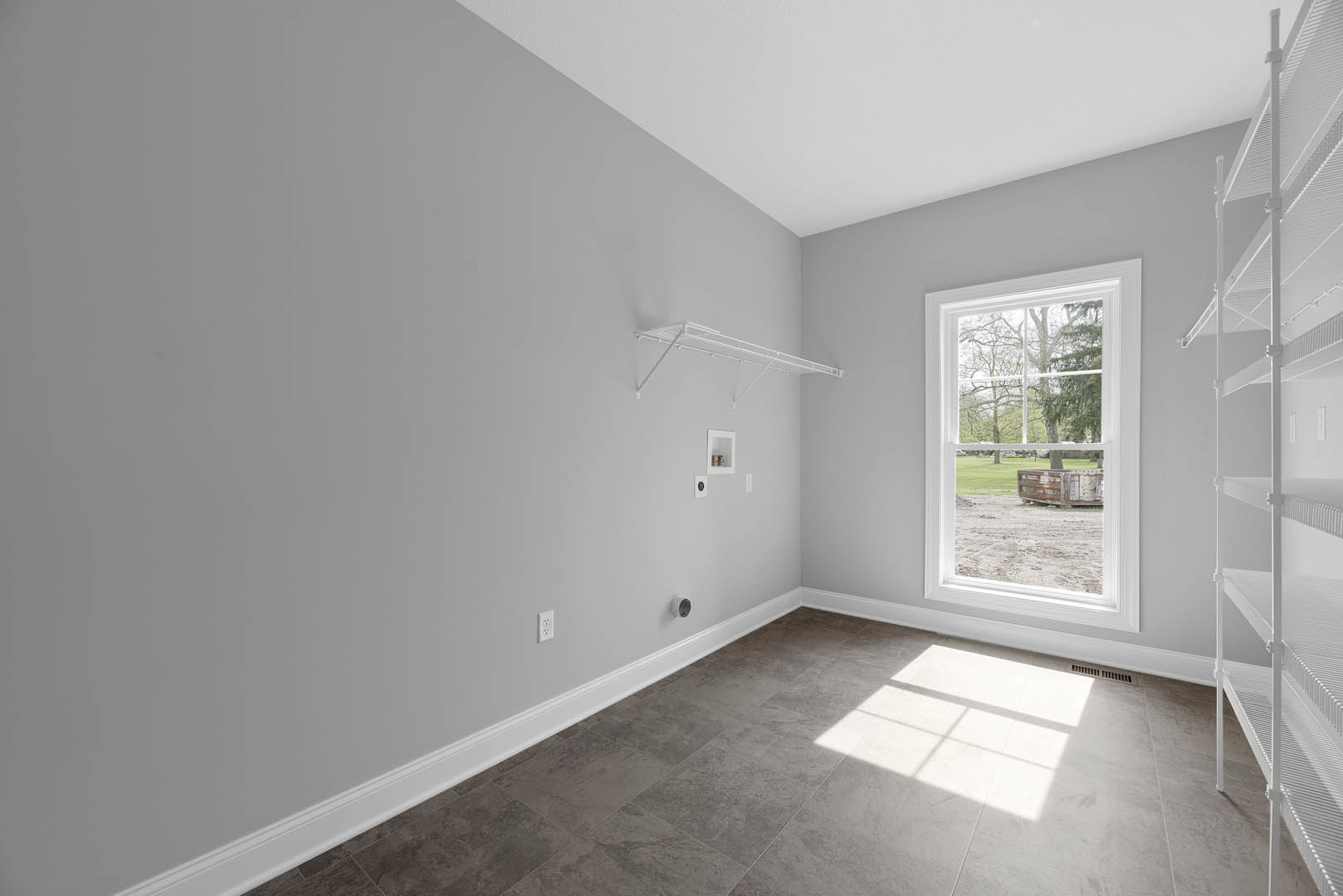 Grey tile floor with white baseboards, white floating shelf mounted on plaster wall, large window overlooking grassy yard with tree.