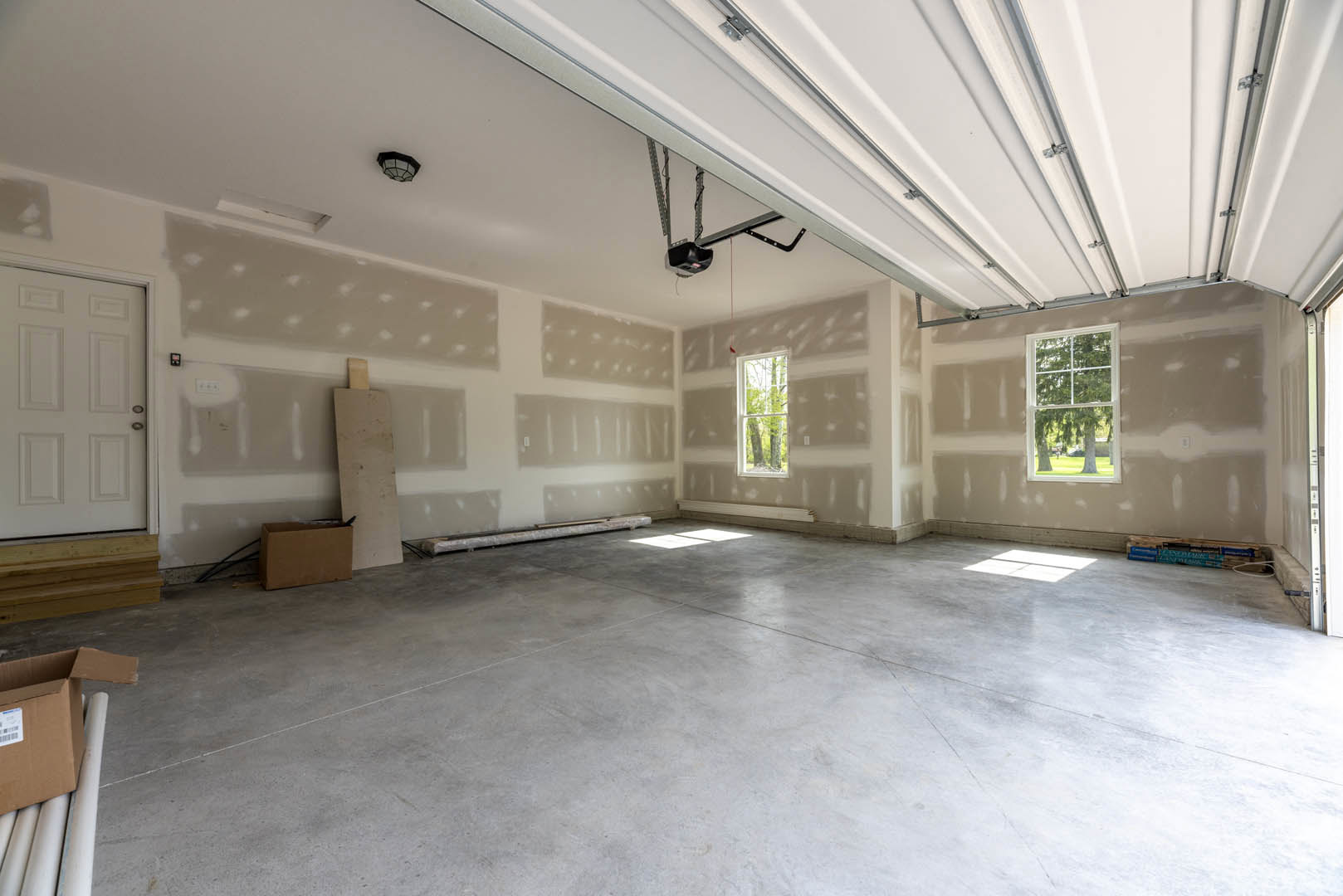Concrete floor with sunlight streaming through a window, white plaster wall, white ceiling with exposed metal rails, white door with silver handle, two stacked cardboard boxes