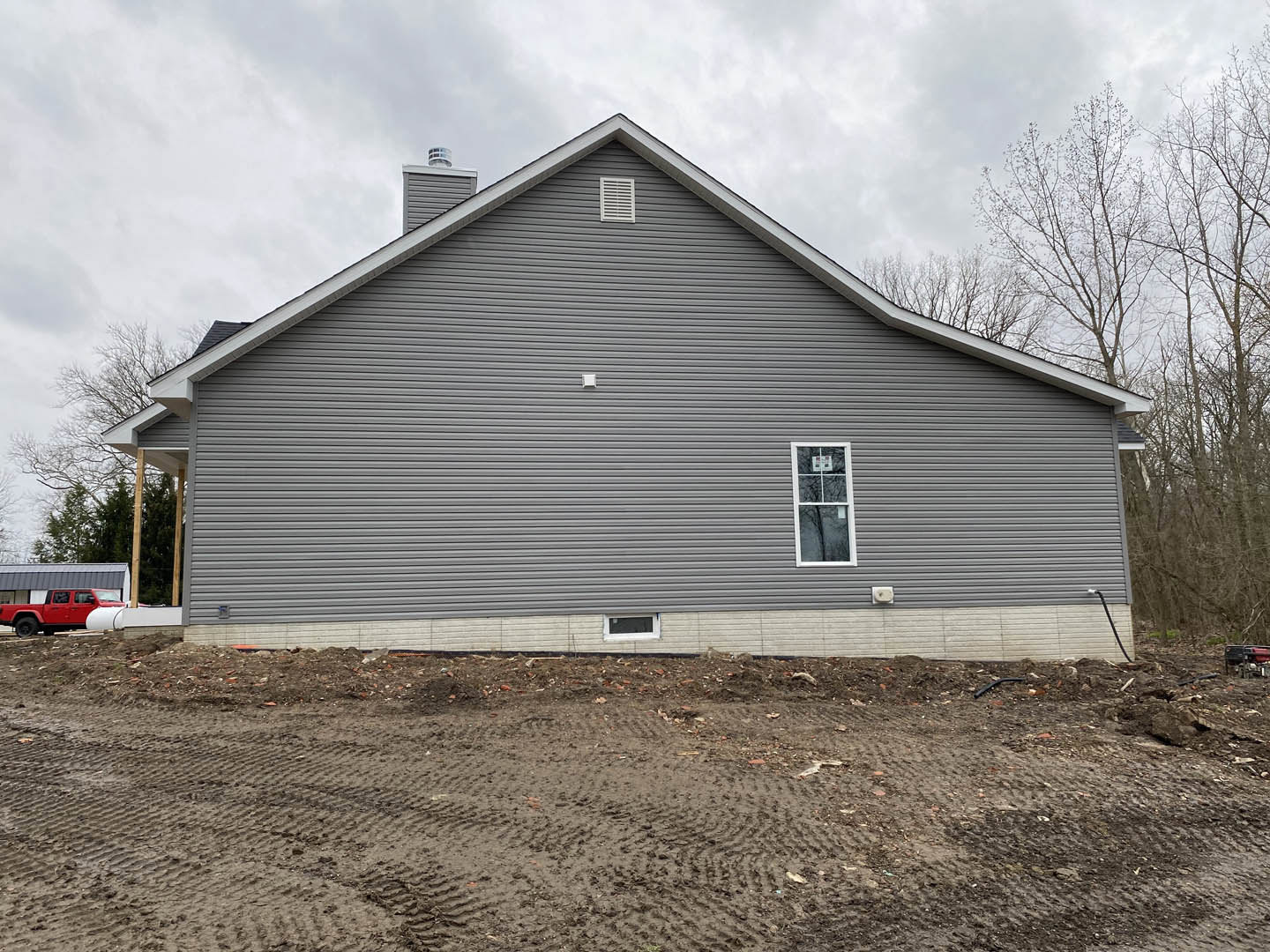 Light gray siding and white trim on a single-story house, basketball hoop mounted in a window, dirt yard with tire tracks, red pickup truck parked beside, mature trees in the