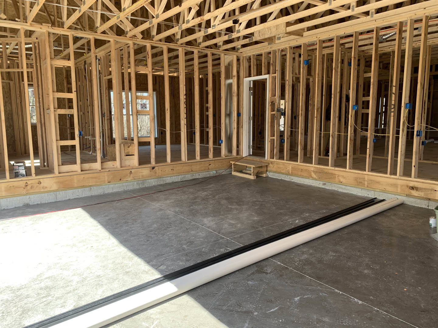 Concrete floor with grey tiles, exposed wood framing, roof truss, large window, wooden table with red rope, close-up of door, construction materials visible throughout interior
