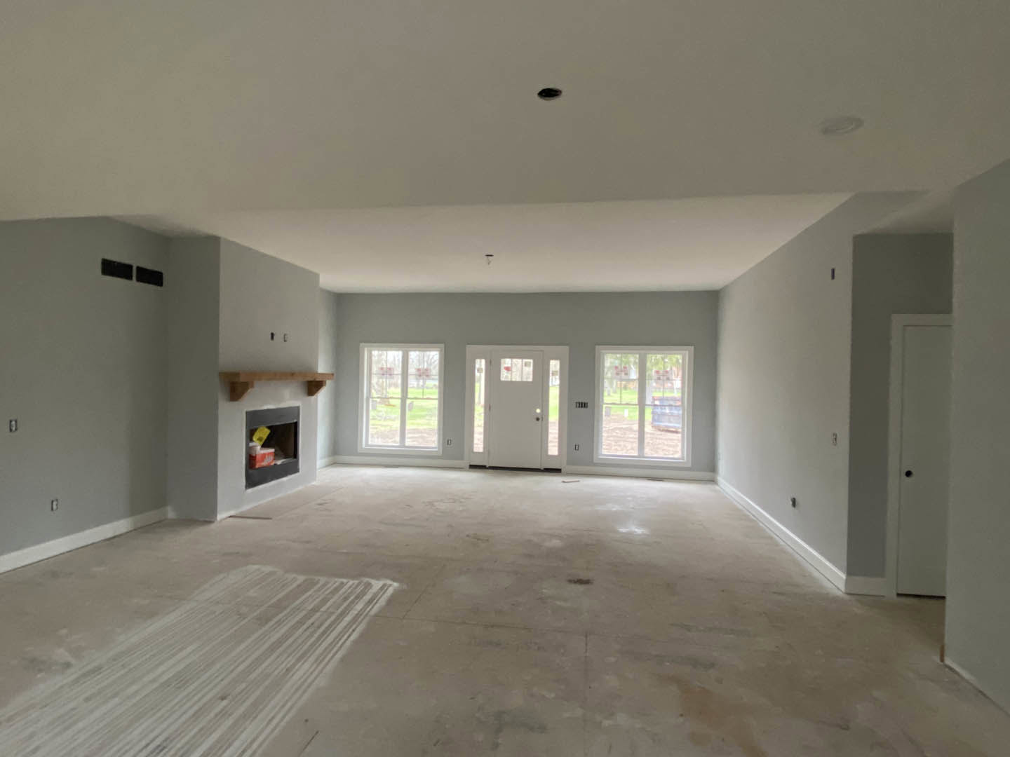 Living room with plaster walls, wood laminate flooring, white fireplace surround, glass-paneled door, and large windows overlooking grass and trees