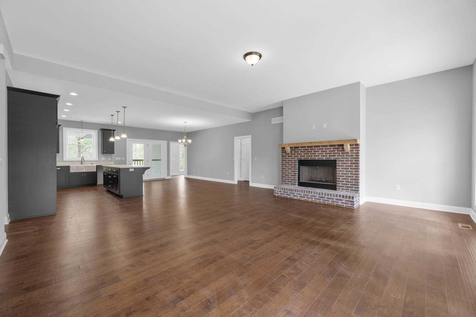 Spacious open living area with hardwood floors, stone fireplace with glass door, modern ceiling light fixture, kitchen island with marble countertop, person near a grey accent wall