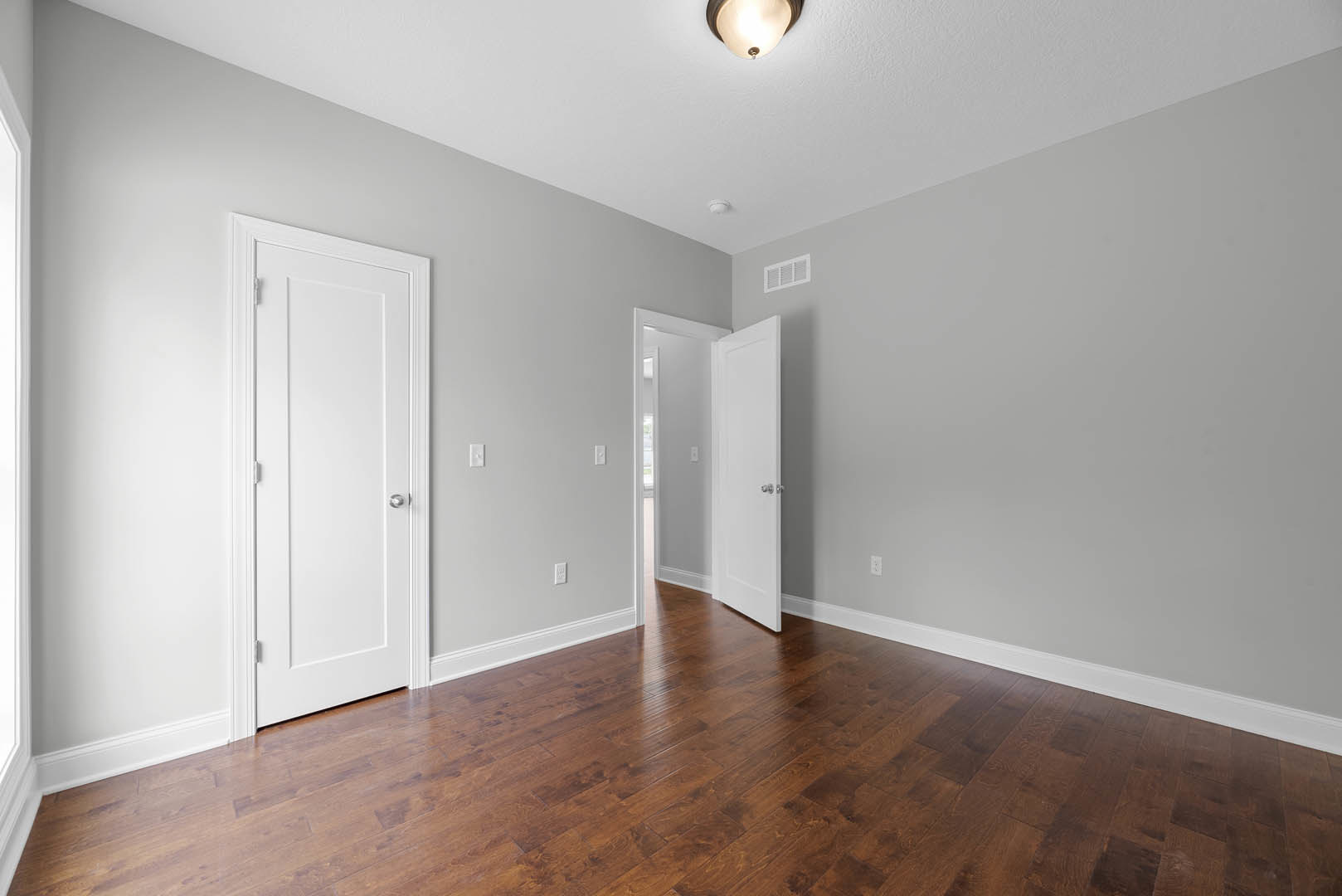 White paneled door with silver knob open onto wood laminate flooring, white wall with vent, ceiling light fixture visible