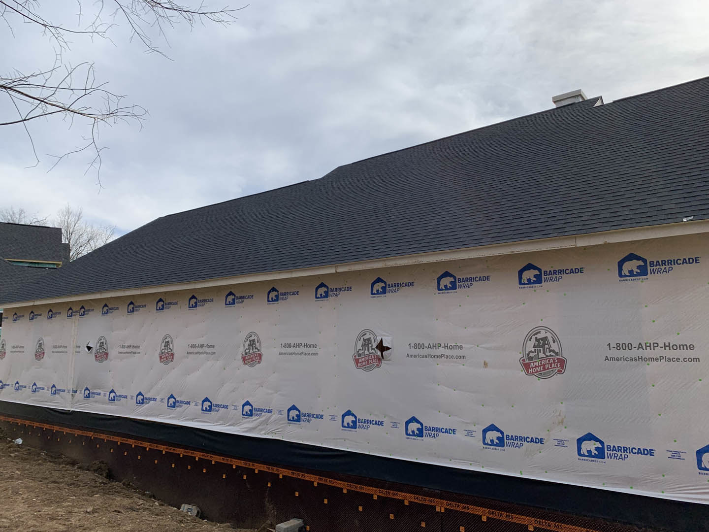 Modern custom home exterior with white tarp covering one side, blue and white logo displayed on surface, pitched roof, surrounding trees, and cloudy sky.