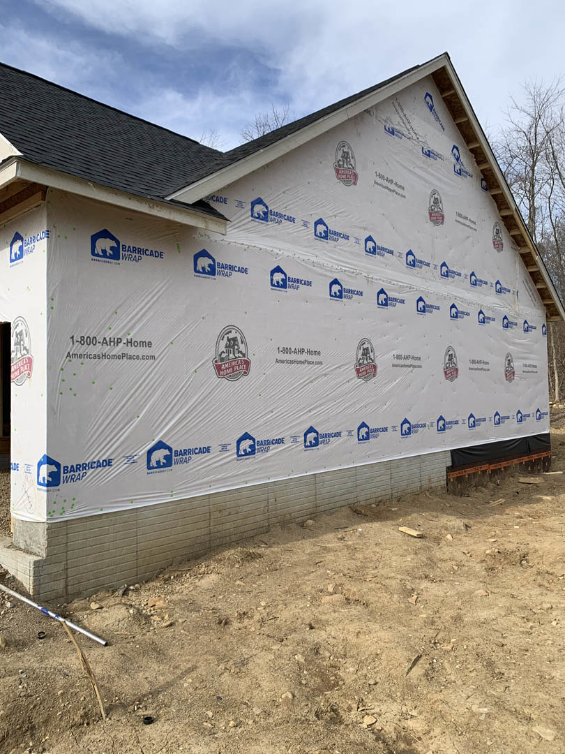 White siding covers the side of a custom home, with a dirt yard in front, a white sign featuring a bear logo, and a metal pole with blue and white stripes on the ground.