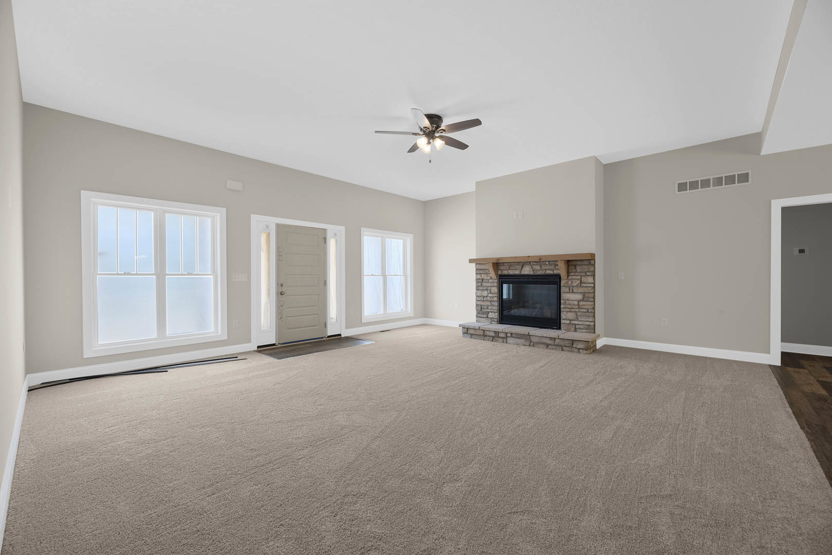 Living room with carpeted floor, stone fireplace with glass door, ceiling fan with lights, plaster walls, and large window.