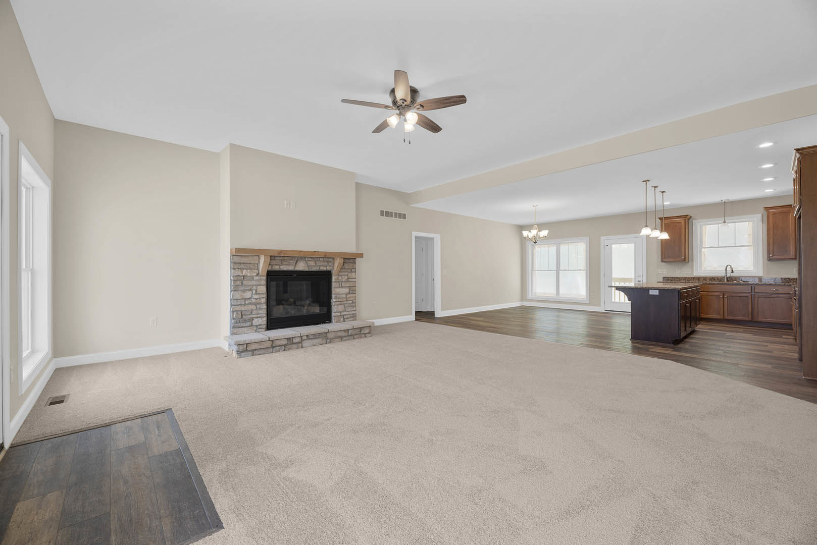 Open living room with wooden floors, marble kitchen island, ceiling fan with lights, glass-front fireplace, and plaster walls.