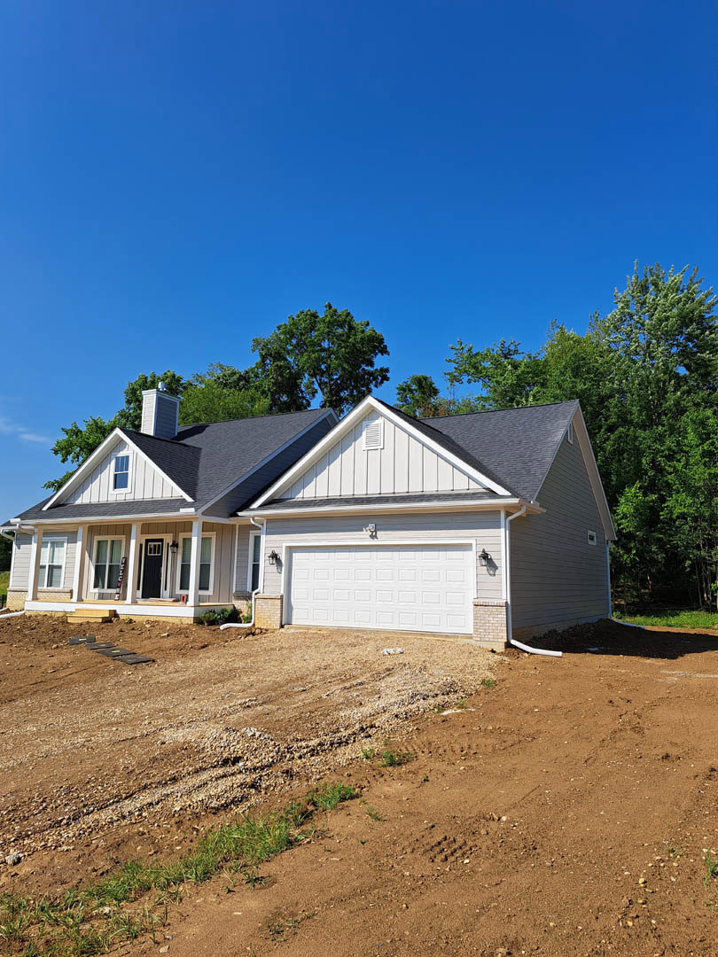Two-story house with white garage door, dirt driveway bordered by grass, leafy trees in background, and a sloped dirt hill beside the property