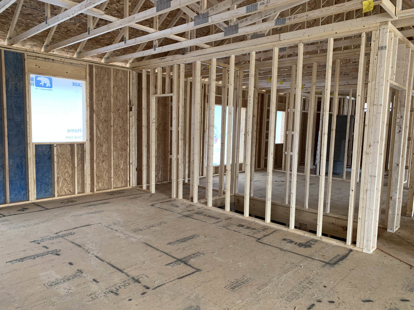Wood-framed room under construction with exposed ceiling beams, unfinished floor marked in black, partial blue wall, and sandy ground showing tire tracks.