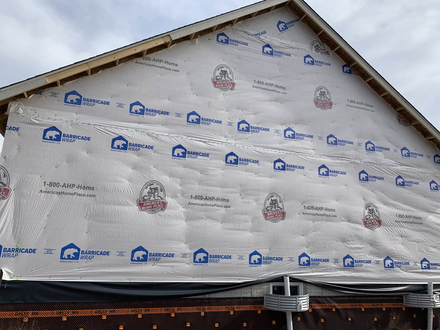 Two-story home exterior wrapped in translucent plastic sheeting, white pipe extending from window, blue square with white bear logo on box near entrance, cloudy sky overhead.