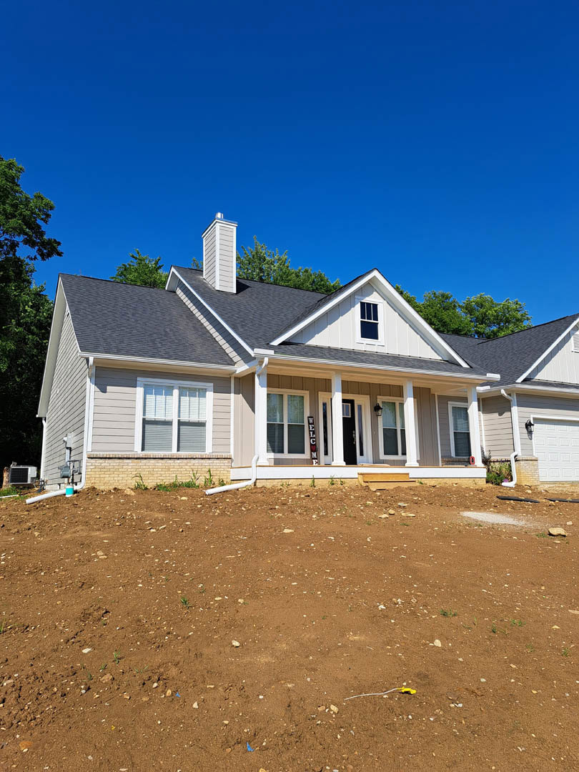 White house with horizontal siding, welcome sign near entry, dirt hill and field in foreground, hose on ground, large windows with closed blinds, clear blue sky above