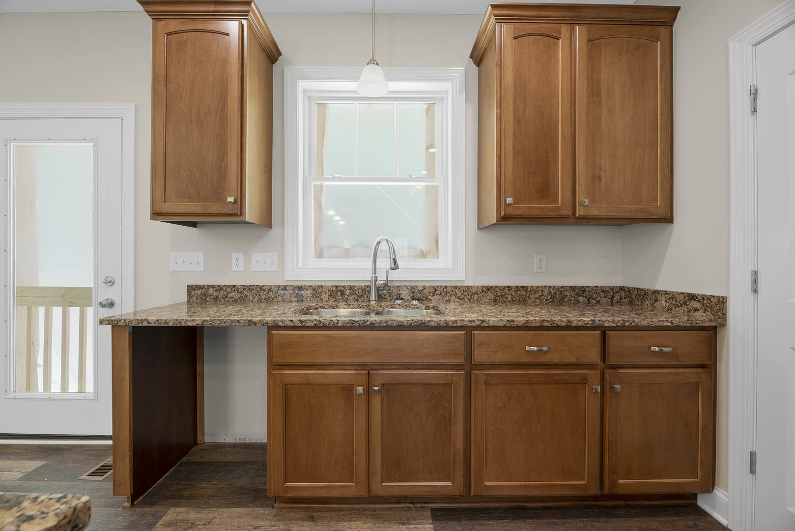 Granite countertops with wooden cabinets, stainless steel sink beneath a white-framed window, glass door, ceiling light fixture, and modern faucet in a residential kitchen.