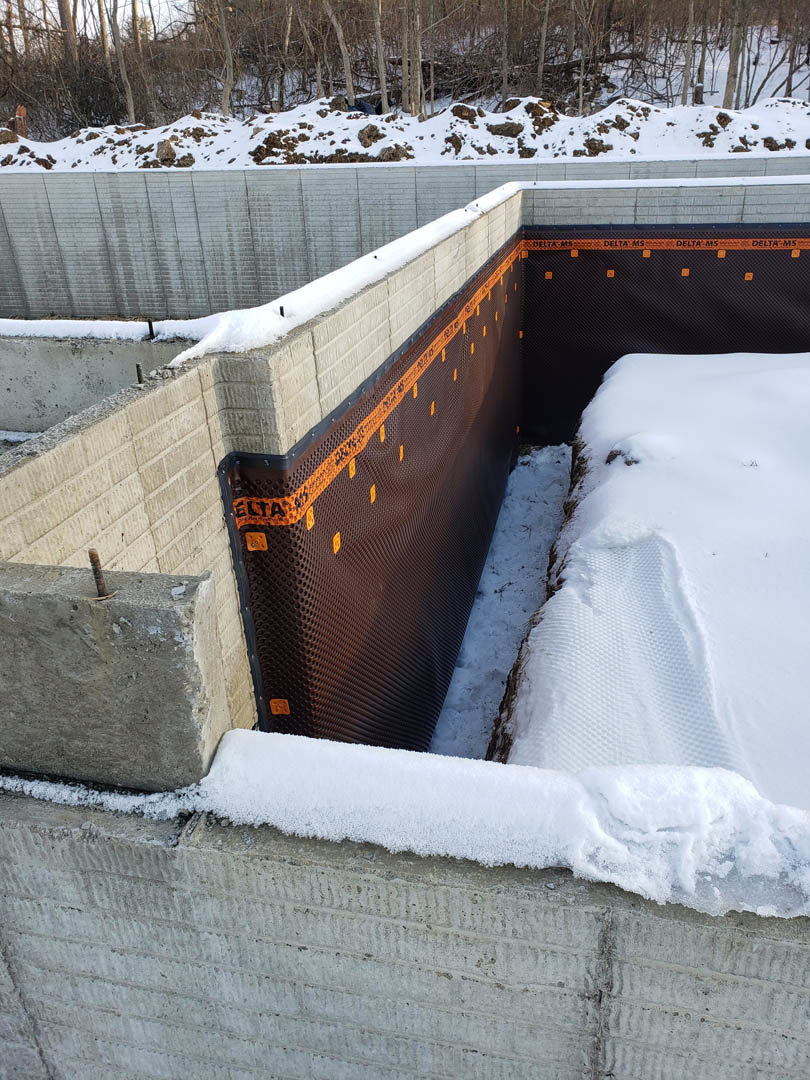 Snow-covered foundation with brown and black mesh protective layer, surrounded by snowy forest and trees