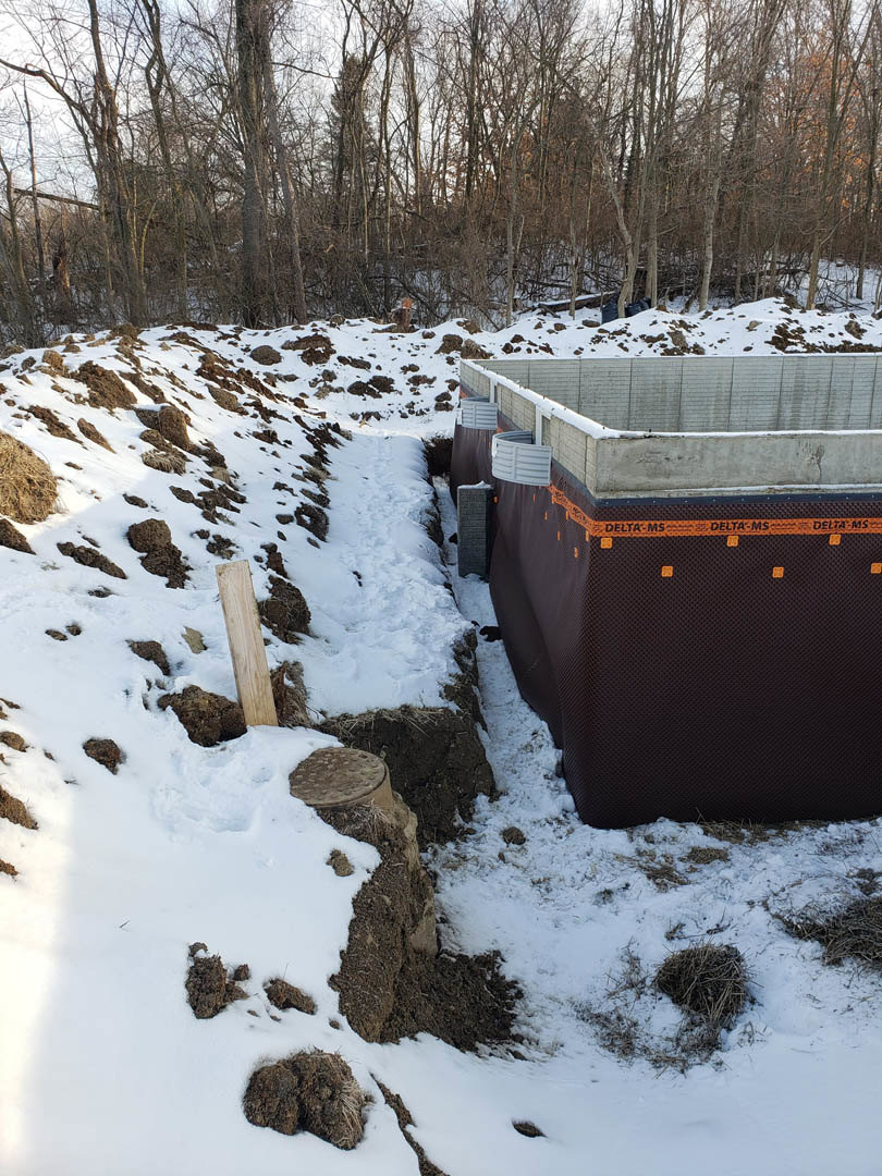 Concrete foundation with black cover surrounded by snow, forest trees in background, wooden post and pile of dirt in foreground, round brown object with dots on snowy ground