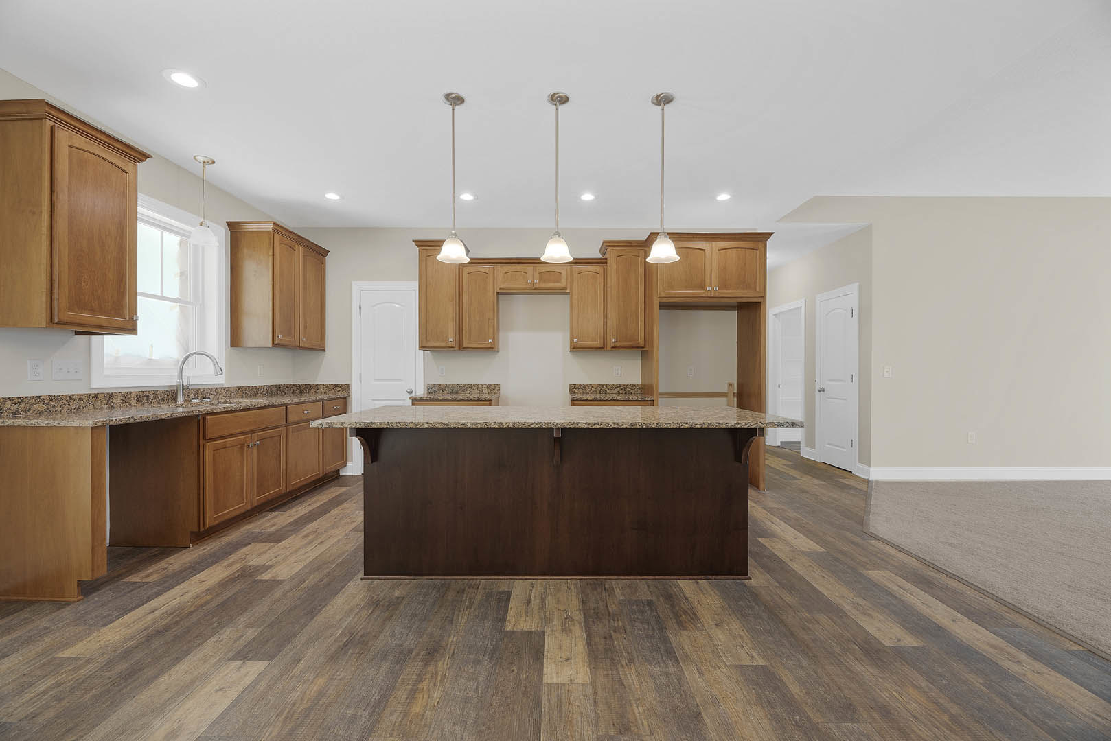 Spacious kitchen featuring a large central island with stone countertop, white cabinetry, stainless steel faucet, pendant light fixtures, and hardwood flooring.