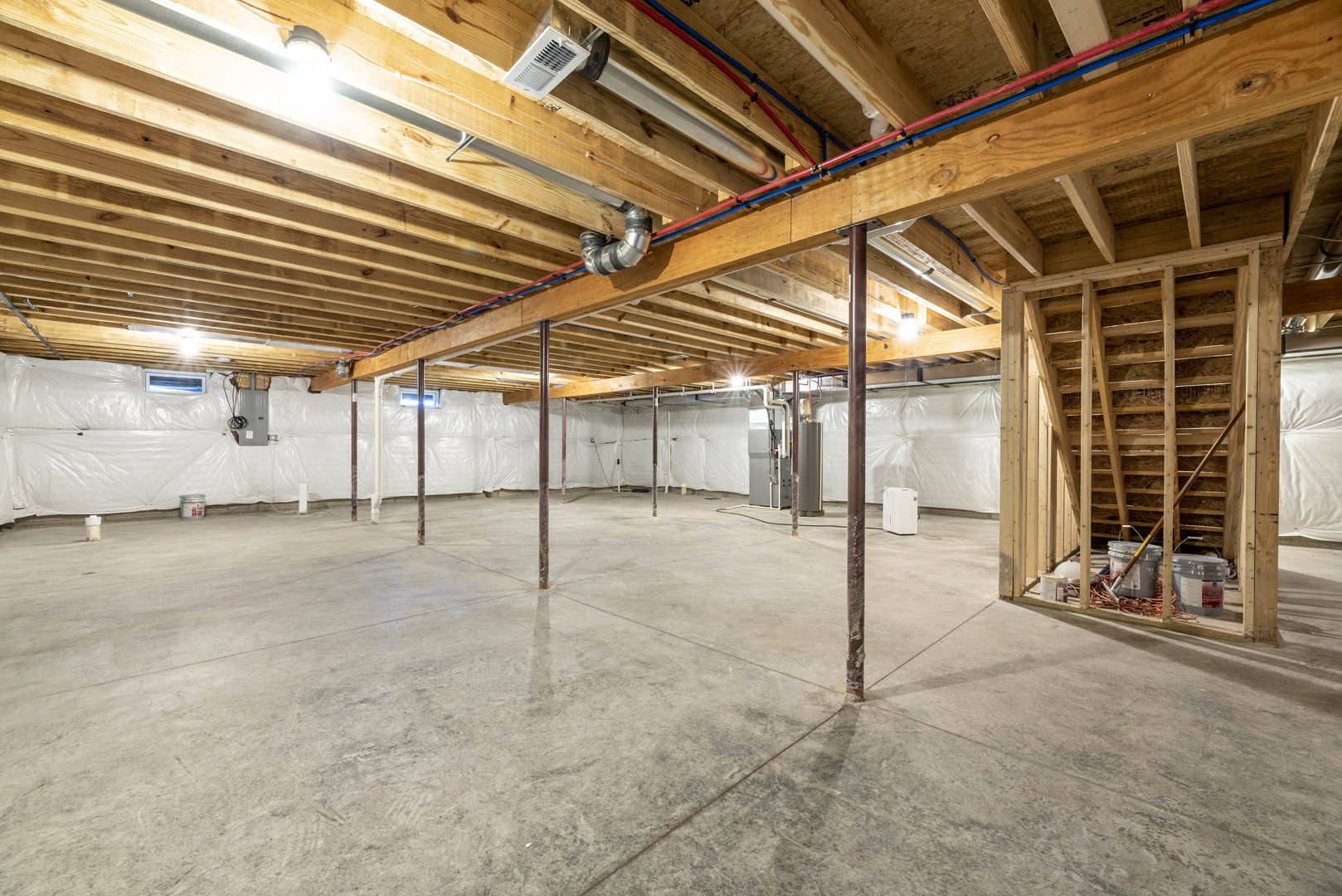 Basement with exposed wood beam ceiling, concrete floor, white vent on wood surface, metal pipe with black rubber bands, and a white box on the floor.