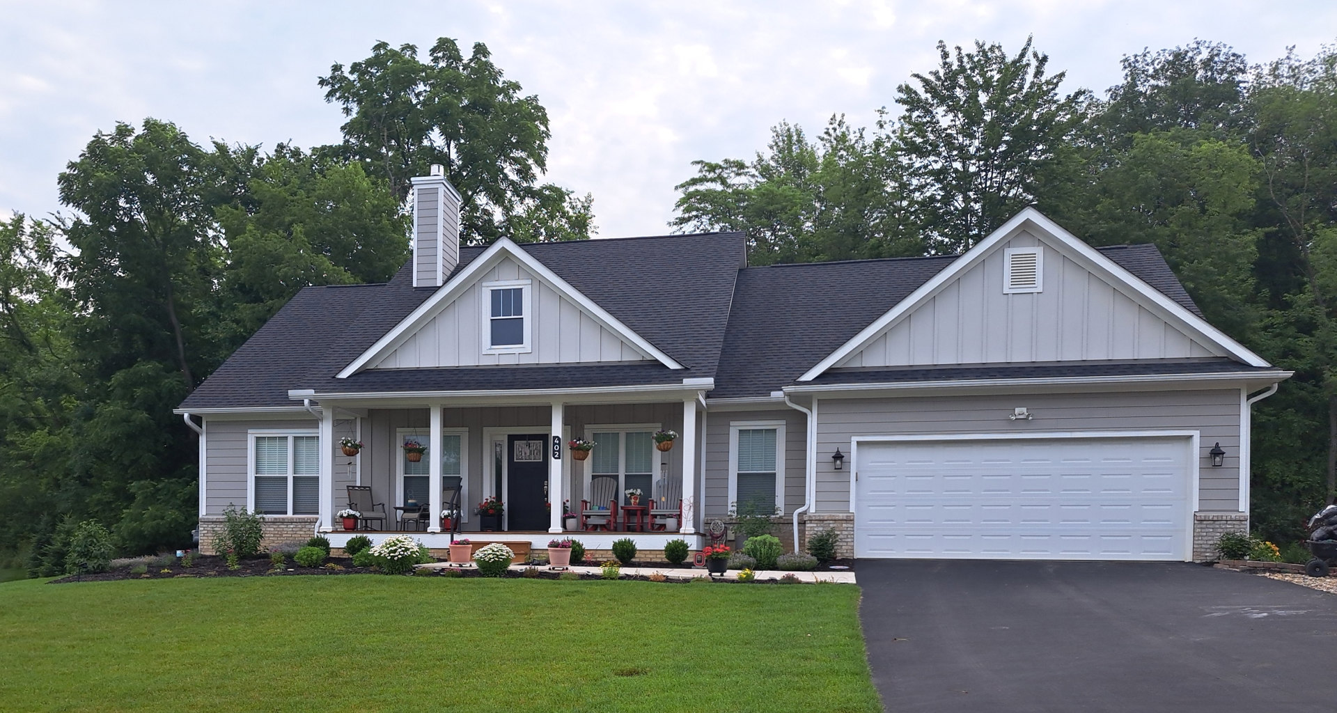 Two-story house with white siding, white-framed windows, and a metal-framed garage door, surrounded by green lawn, bushes with white flowers, and a paved driveway