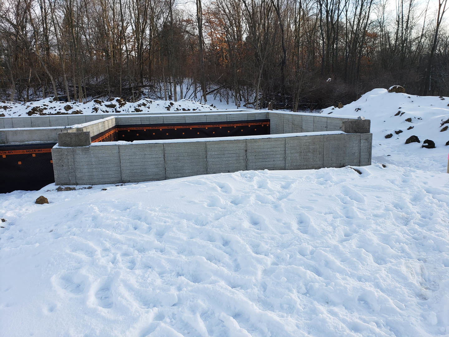 Concrete foundation surrounded by deep snow, man standing beside snow-covered wall, snowy forest and hills with footprints and rocks in background