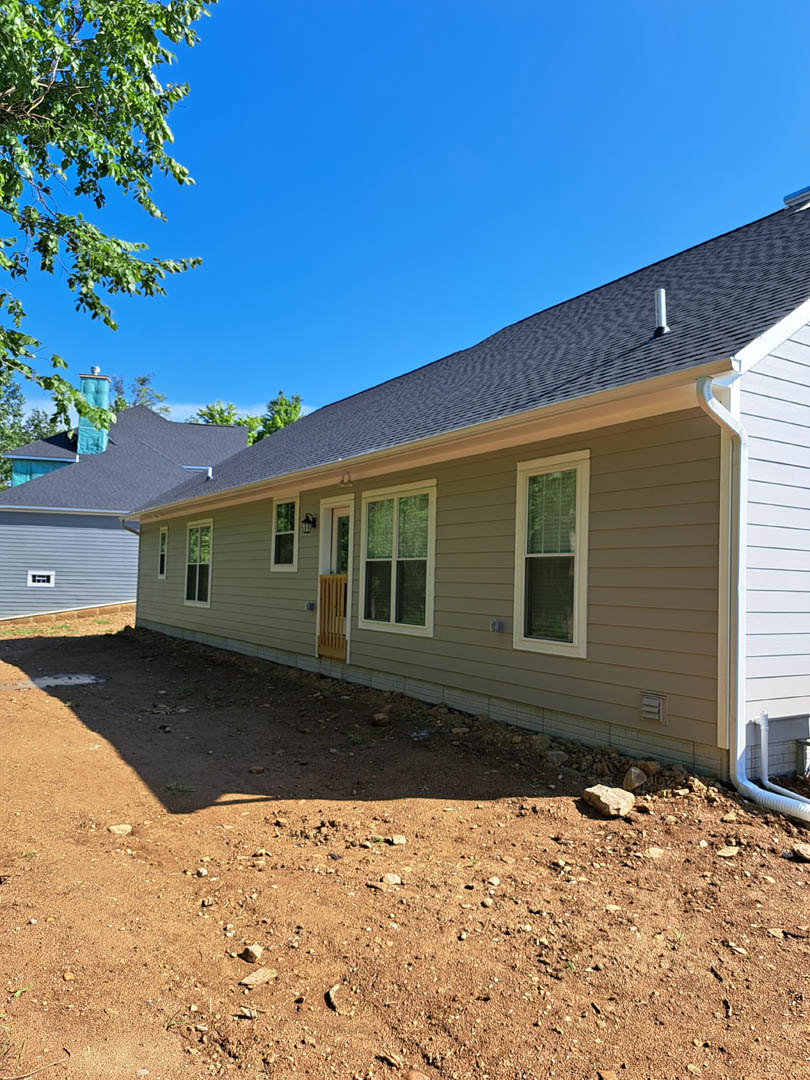 Two-story home with white-framed windows, light siding, dirt and rocks in the yard, mature trees and green leaves in the background