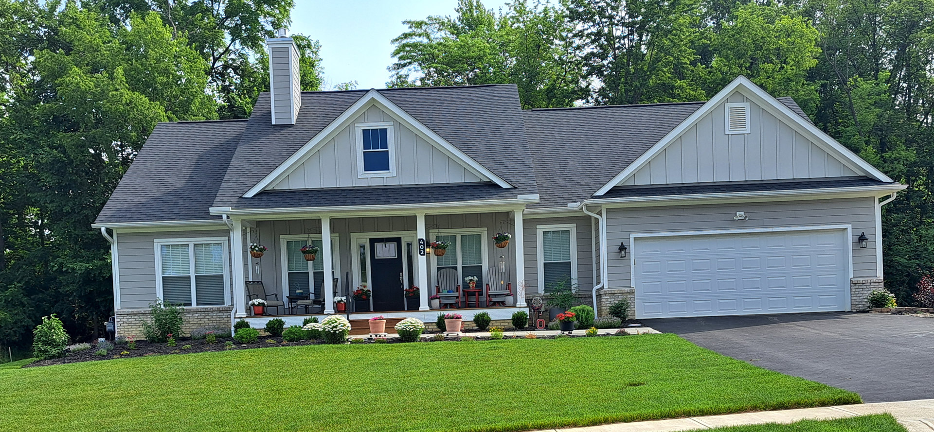 Two-story house with white siding, large windows, covered porch, green lawn, mature trees, and landscaped flower beds