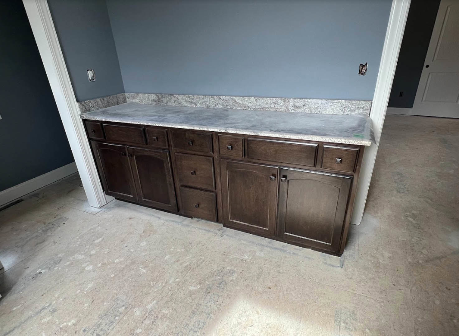 Brown wood kitchen cabinets with marble countertop, white wall, and drawers, adjacent to a white door with a hole.