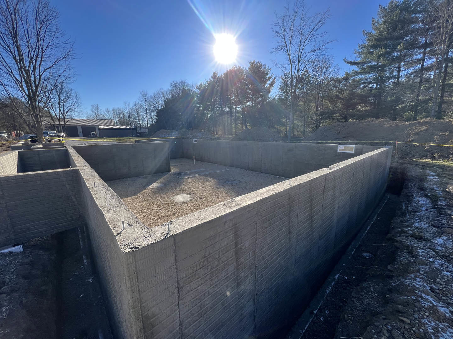 Concrete foundation set on bare ground with leafless trees and blue sky in the background, sunlight filtering through branches.