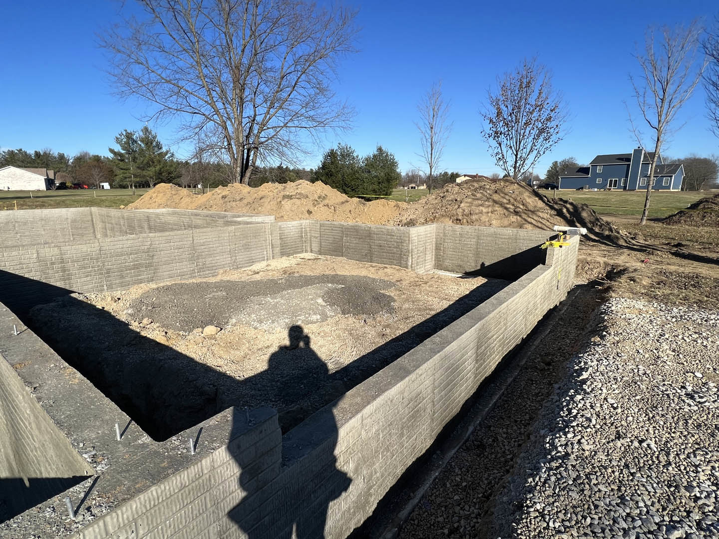 Concrete foundation surrounded by dirt, leafless tree and group of trees under blue sky, shadow of person holding phone cast on ground, blue house with chimney in background