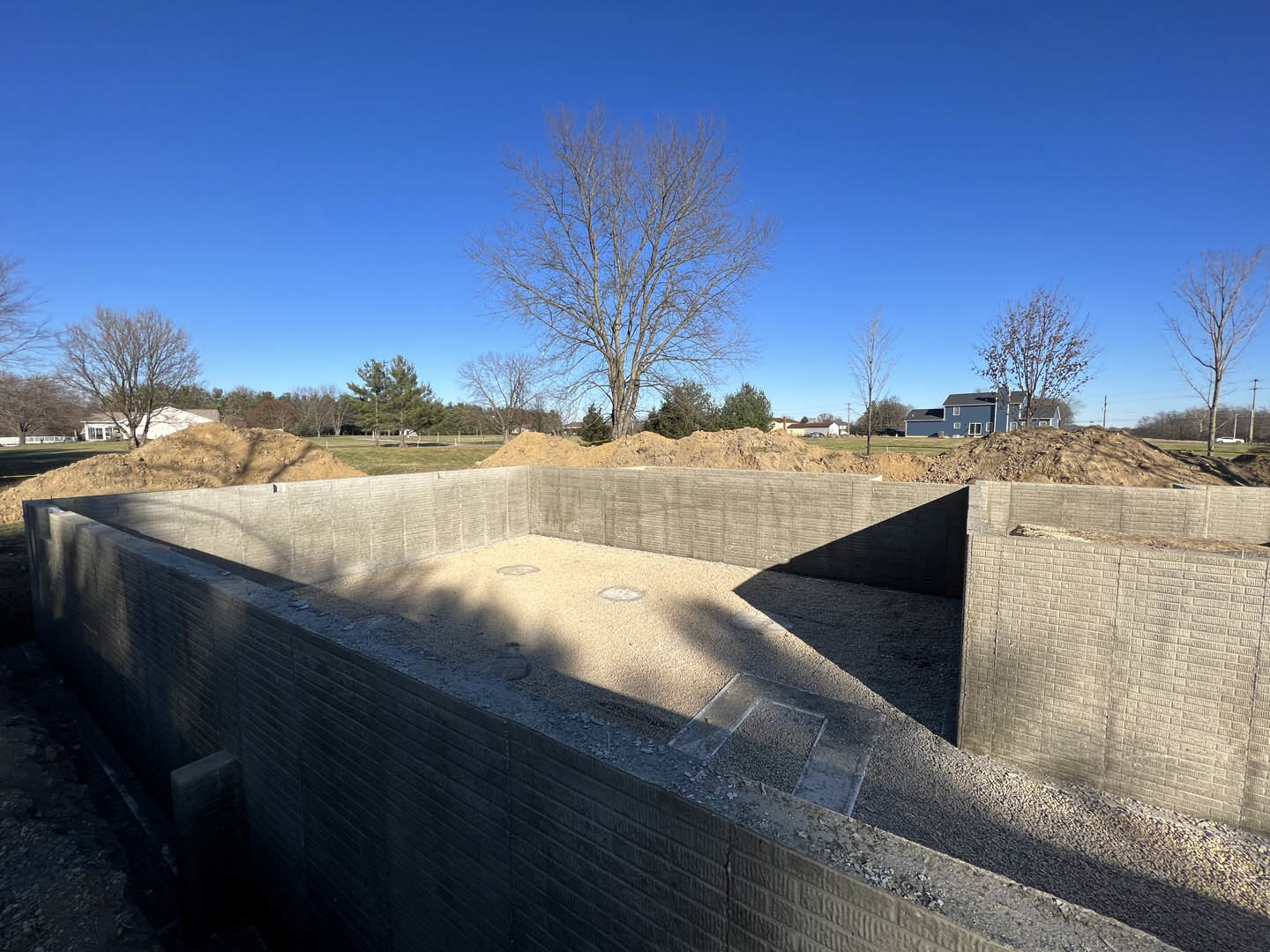Concrete foundation surrounded by dirt, leafless trees, and a pile of gravel in the background