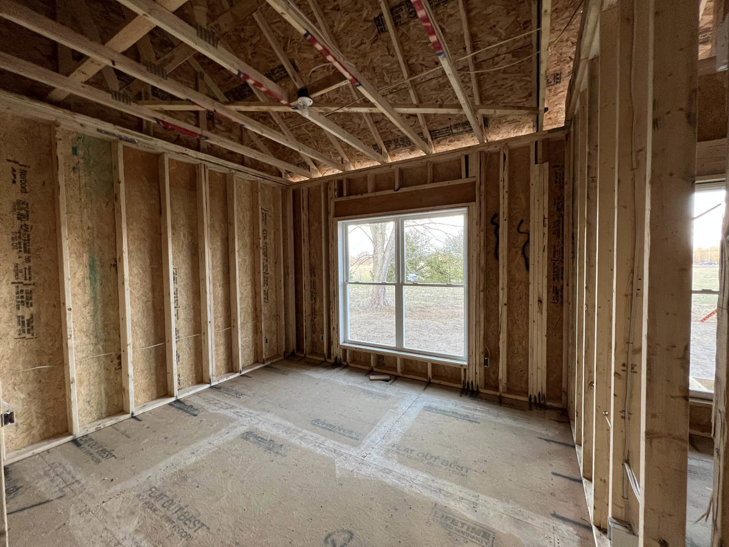 Bright room featuring exposed wood ceiling beams, large window overlooking leafy trees, light-colored walls, and unfinished flooring with visible opening.