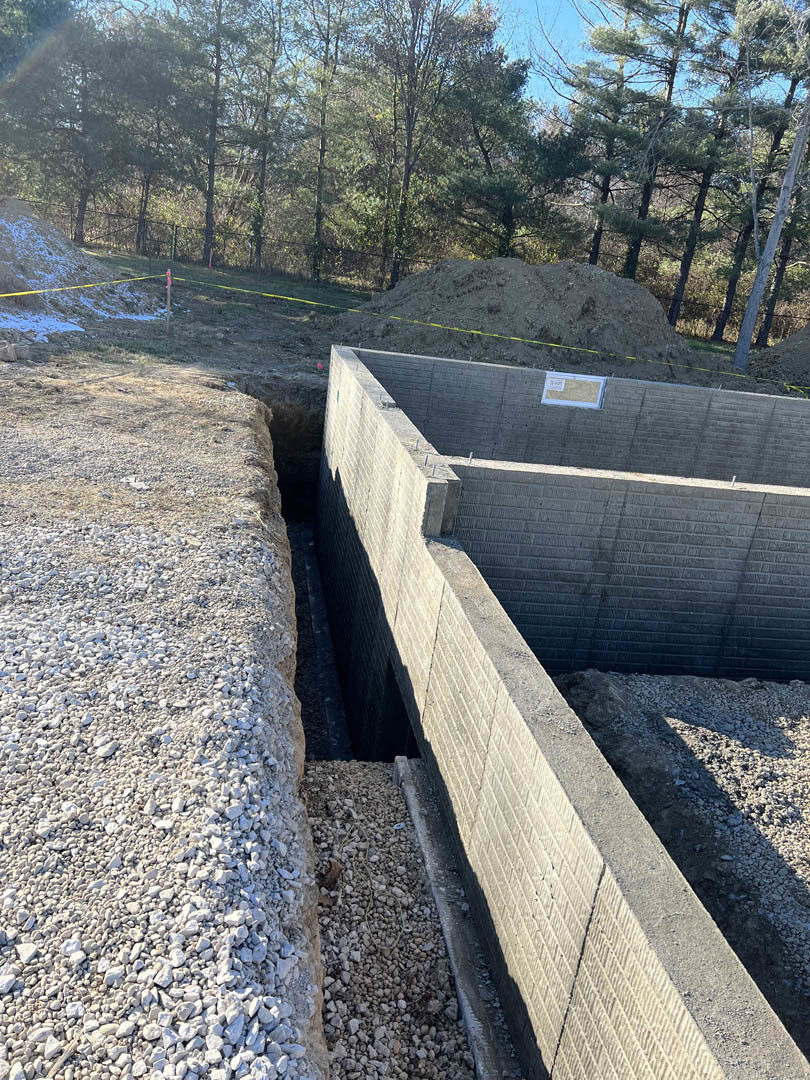 Concrete retaining wall with a circular hole in the ground, piles of white rocks and dirt, yellow caution tape stretched across a hill, trees and plants in the background.