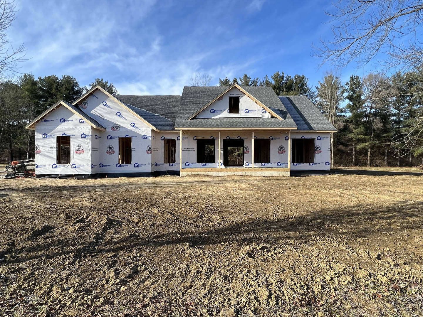 Partially built house with exposed framing, black-framed windows, white construction materials on dirt ground, blue sky and scattered clouds, trees in background