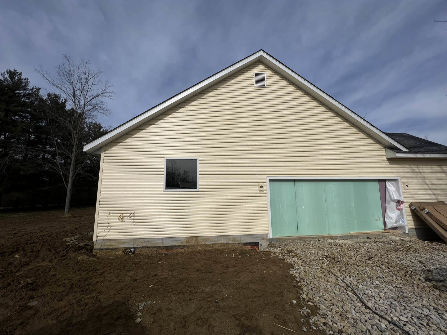 Modern house with attached garage, green siding with white trim, large window reflecting nearby trees, tree beside the entry, white plastic bag with red logo on driveway