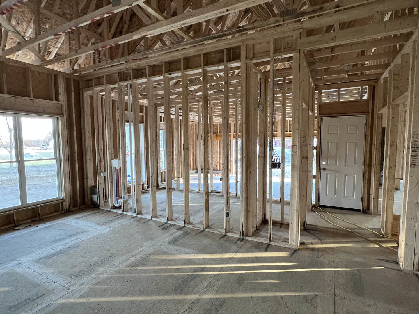 White door with silver doorknob set in a wall, wood ceiling beams overhead, large window offering view of grassy field and trees, concrete floor with wooden framing visible.