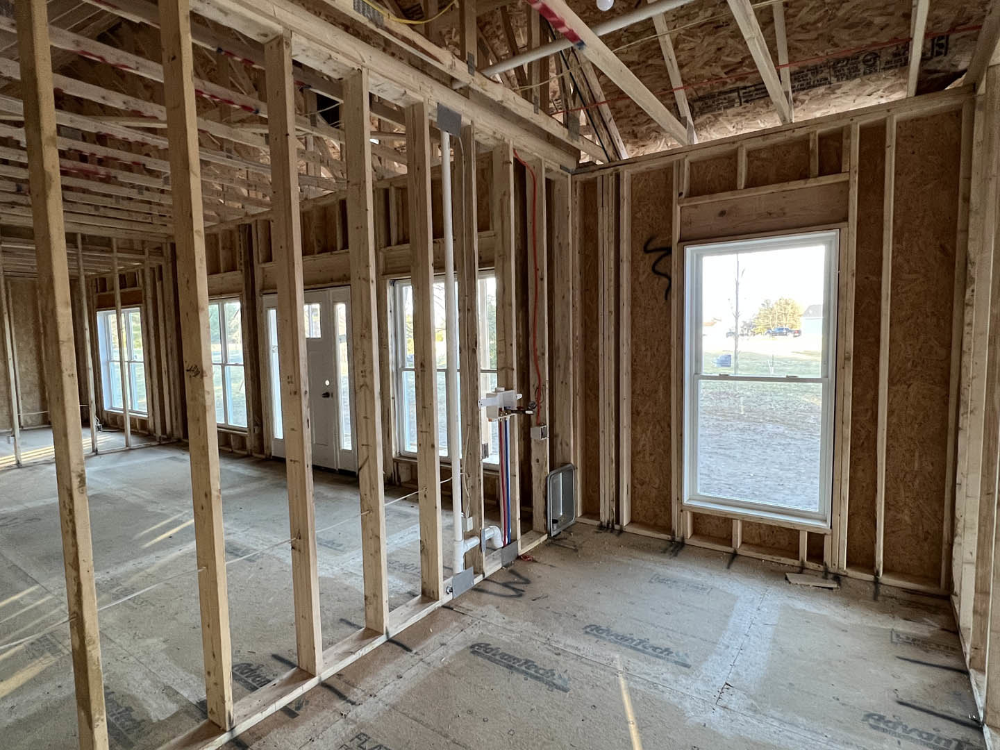 Wood-framed room under construction with exposed beams, unfinished floor, door, and window overlooking a field and trees