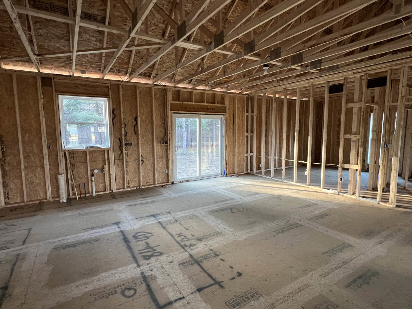 Unfinished room with exposed wooden roof trusses, concrete floor marked with black writing, large sliding glass door, and window overlooking a tree.