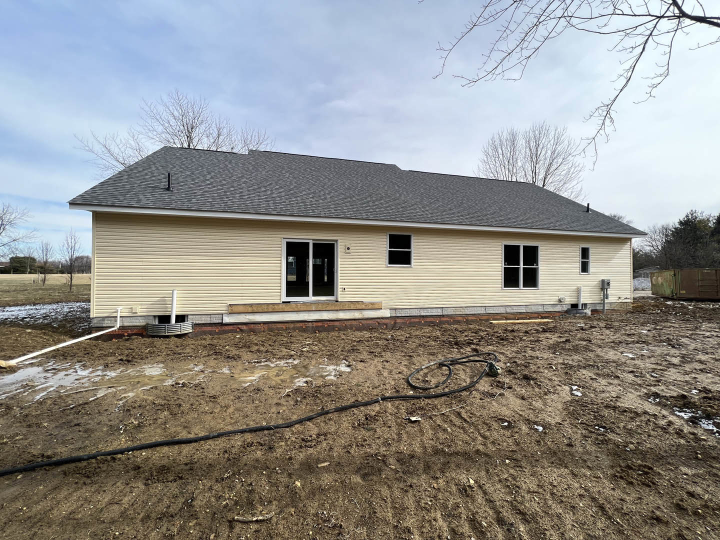 Modern house with light-colored siding, open sliding door, white-framed windows, dirt yard, garden hose on ground, and cloudy sky overhead
