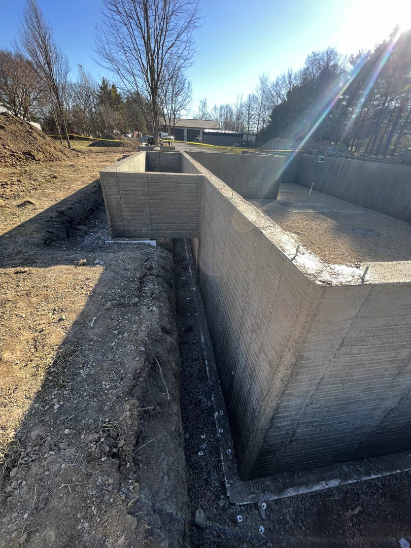 Concrete foundation with a large hole in the ground, leafless tree and blue sky in background, dirt and rocks scattered around, brick wall and building visible behind.