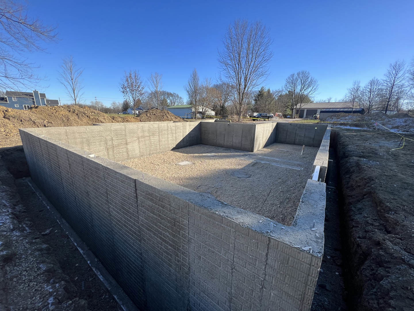 Concrete foundation bordered by gravel, exposed dirt in the center, leafless trees in the background, partial view of a building with a window and roof.