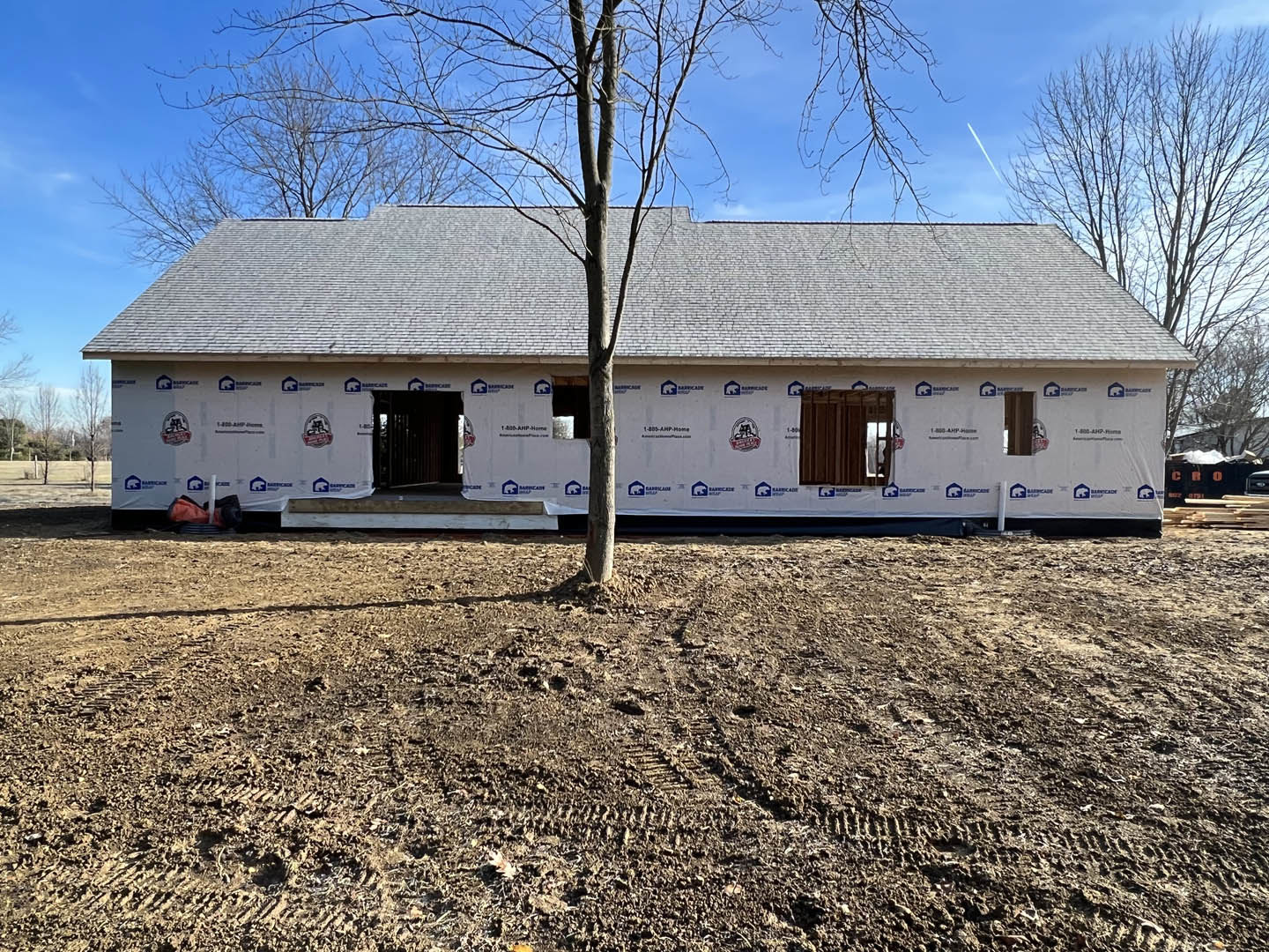 Framed house under construction with exposed wood, dirt lot, and leafy tree in the backyard