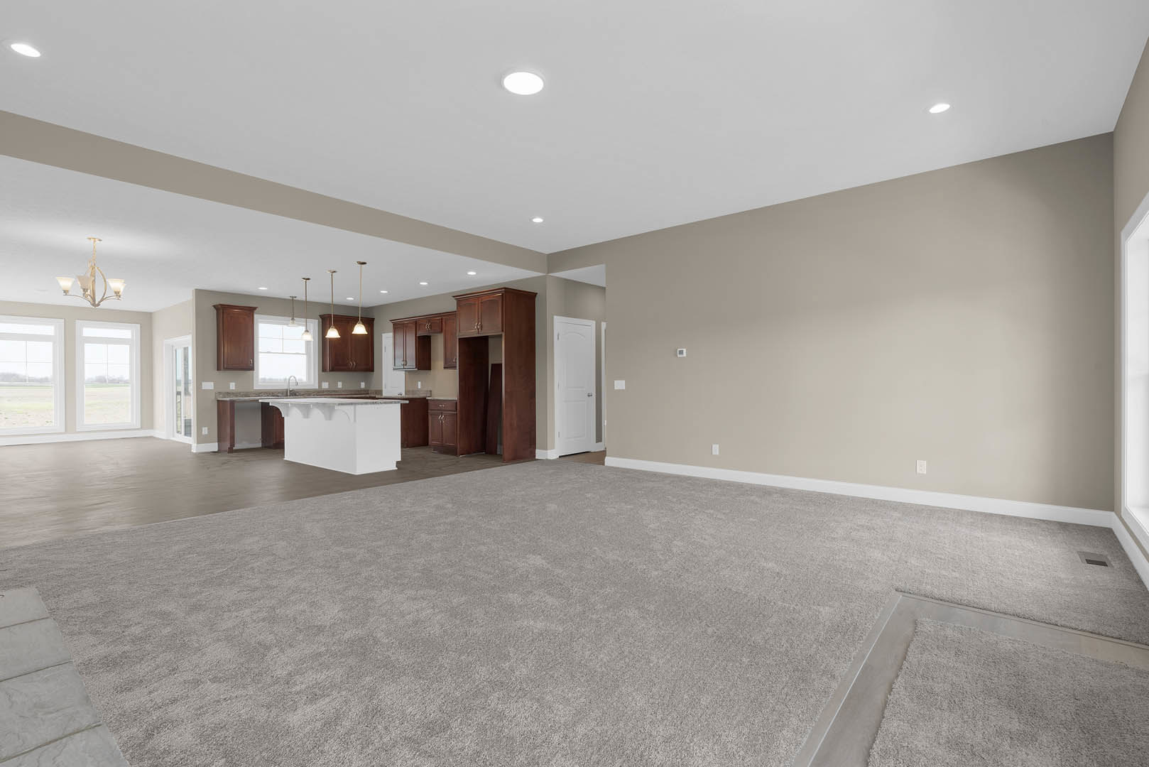 Open living room with beige carpet, adjacent kitchen featuring tile flooring and a bar with a clear countertop, white door with silver handle, ceiling light fixture with three
