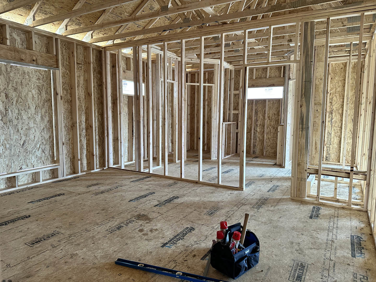 Wood-framed room under construction with exposed beams, carpeted floor, bag of tools, blue level, red and silver spray can, window displaying a sign, and white screen with blue