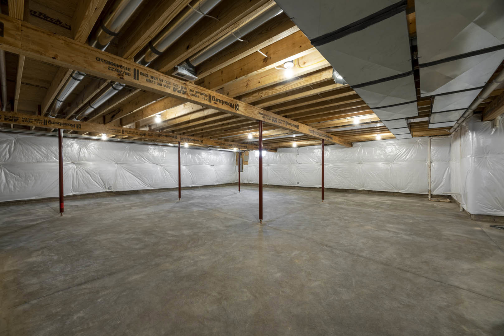 Concrete floor with central metal pole, exposed wooden ceiling beams, white walls, visible pipes, and metal ceiling panels.
