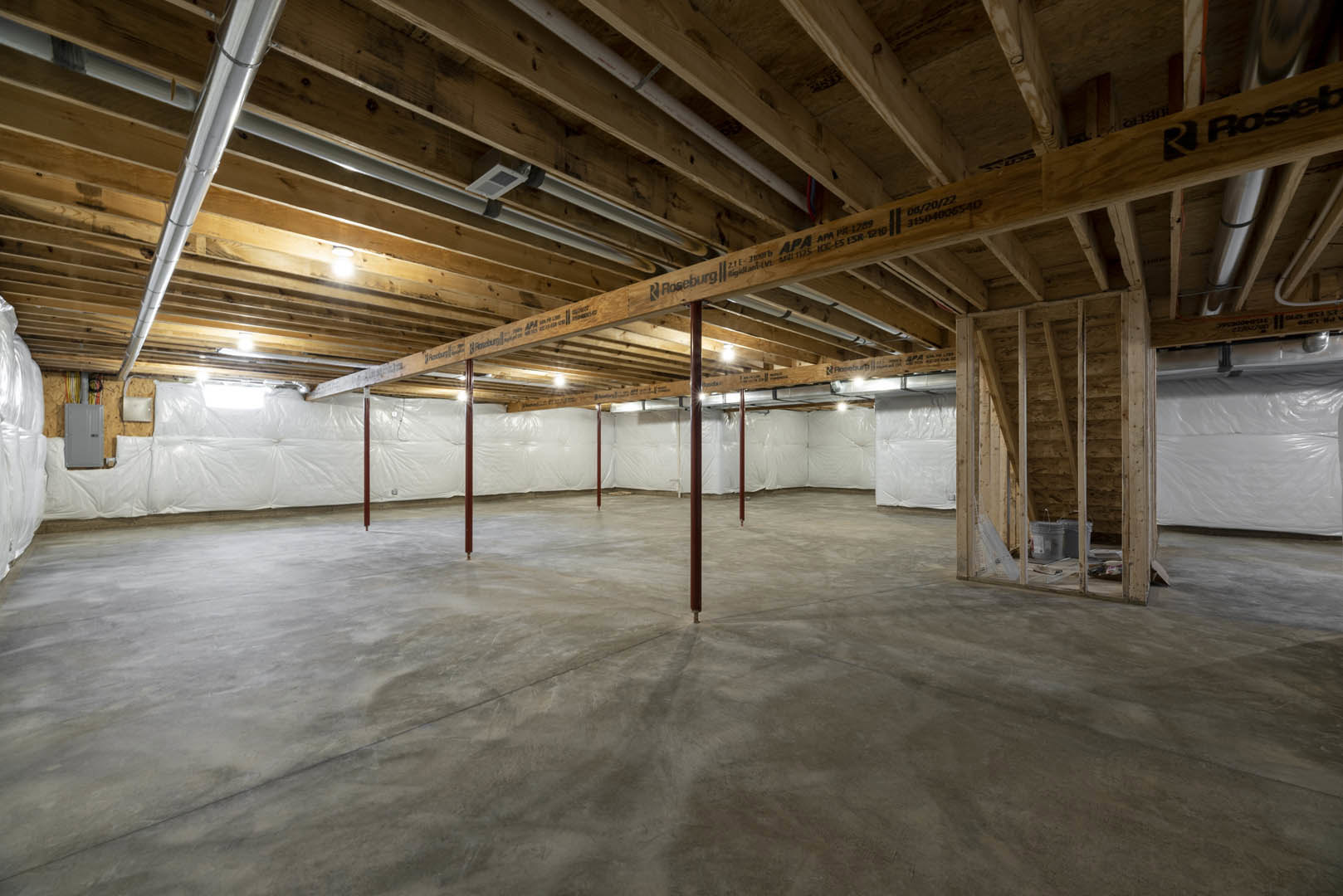 Exposed wooden ceiling beams, white ceiling and walls, concrete floor with metal poles, grey door with black handle, white plastic bag on bed, white towel on white surface
