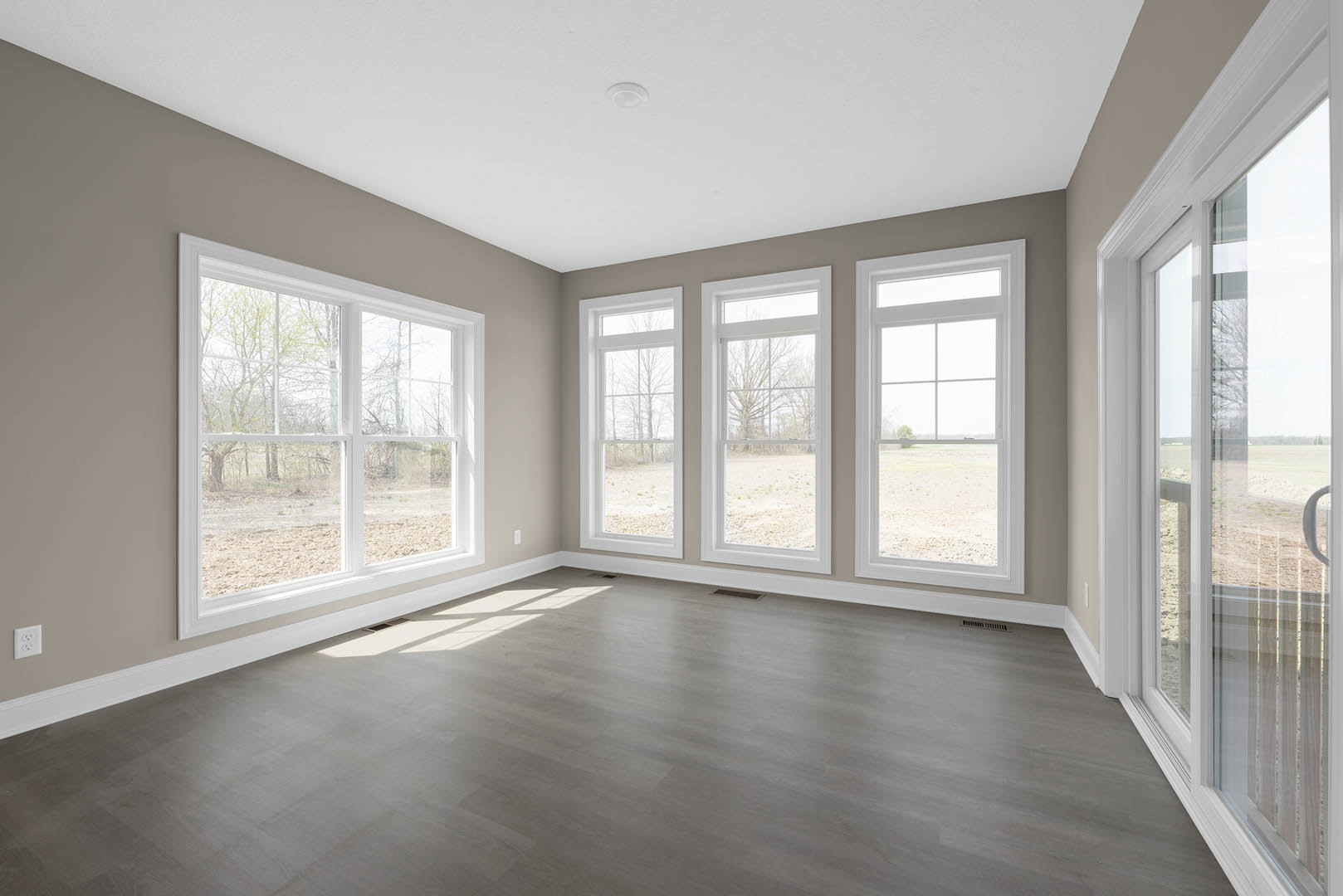 Spacious room featuring multiple large windows, wood flooring, white plaster walls, and views of trees and open fields outside